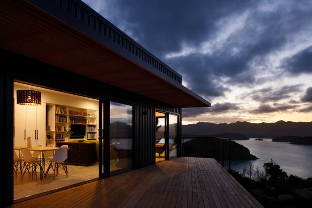 A modern dark-clad coastal home at dusk, viewed from a generous timber deck overlooking a calm bay and silhouetted hills. Large sliding glass doors open directly to a warmly lit open-plan dining and living area, with timber cabinetry, bookshelves, a pendant light, and dining table visible inside. The projecting upper deck or roof edge creates a strong horizontal cantilevered form.