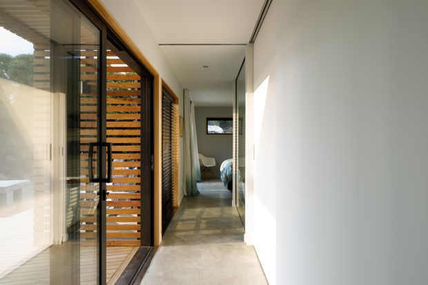 A bright, narrow modern hallway or gallery corridor with polished concrete flooring, plain white walls, and full-height glazing and sliding doors on the left. Warm timber louvres and screens filter sunlight beside the glazing, while a large mirrored wall on the right reflects the corridor and furnishings. A bedroom or sitting area is visible at the far end.