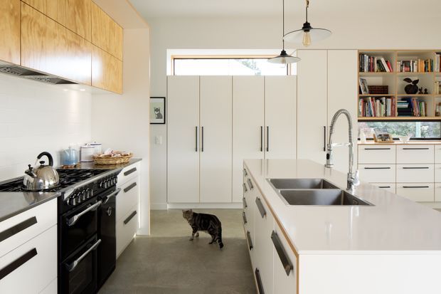 Bright, spacious contemporary kitchen with white flat-panel cabinetry, a large central island with double undermount sinks, integrated dishwasher and seating overhang, a dark freestanding range, timber overhead cabinetry, open shelving, and concrete flooring. Pendant lights and clerestory-style windows provide natural light.