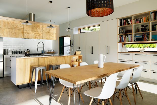 Bright open-plan kitchen and dining area with extensive light timber cabinetry, a large central island, white pendant lights, and a timber dining table surrounded by modern white chairs. Full-height built-in shelving and drawers line the right wall, while a narrow horizontal window brings in natural light. The space has a clean, contemporary look with warm timber accents and polished concrete flooring.