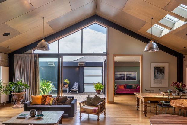 A warm, light-filled open-plan living and dining area with a dramatic pitched timber-lined ceiling, exposed dark structural beams, and high clerestory glazing. The room includes a large sectional sofa, armchair, timber coffee table, pendant lights, indoor plants, and a substantial timber dining table. A glazed courtyard or terrace is visible beyond the living area, with a bedroom or additional sitting room seen through the doorway.