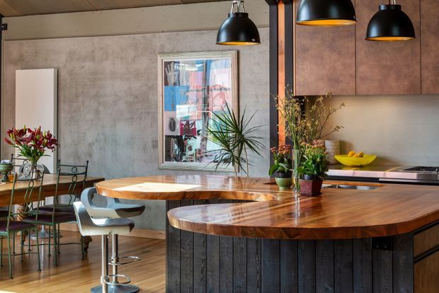 A warm, industrial-style open-plan kitchen and dining area with a large curved island featuring a polished timber benchtop and dark vertical timber cladding. The space includes black pendant lights, dark cabinetry, a concrete-look wall, timber ceiling boards, a round timber dining table, bar stools, plants, and colourful artwork.