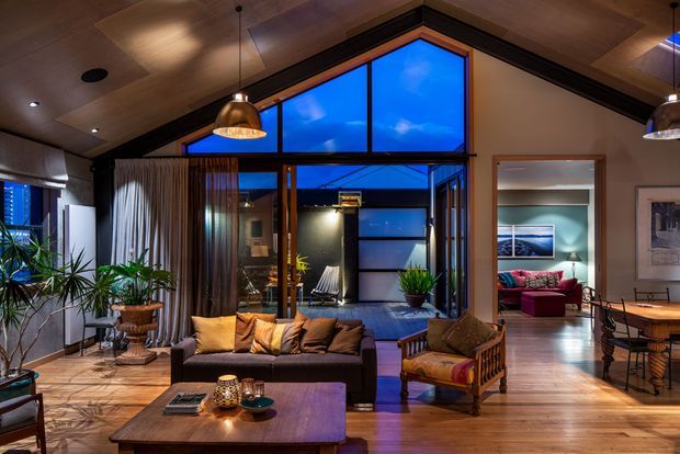 Warm, moody contemporary living room with a large sectional sofa, timber coffee table, armchair, pendant lighting, timber ceiling, and expansive glazed doors and windows opening toward an outdoor area. A dining area and bedroom are visible through the open interior connection, with timber flooring and dark-framed glazing throughout.