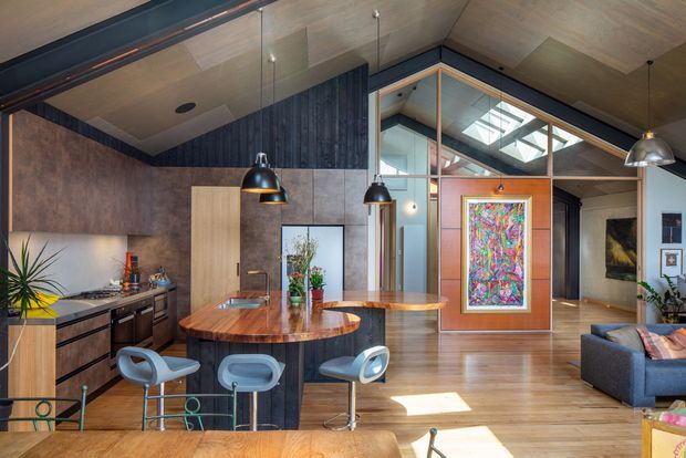 Open-plan contemporary living space combining a kitchen, dining island and lounge. The room features warm timber flooring and a timber-lined pitched ceiling, dark vertical timber wall cladding, wood cabinetry, a sculptural wood island with integrated sink, pendant lighting, exposed structural framing, clerestory/skylight glazing and colourful artwork.