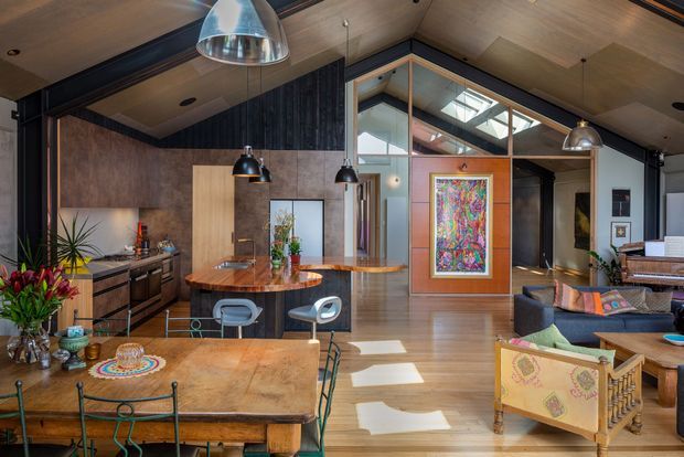 Open-plan contemporary interior combining a kitchen, dining area and living room beneath a high timber-lined ceiling. The space features dark structural framing, warm timber flooring and ceilings, wood-toned cabinetry and island surfaces, pendant lighting, large interior glazing, colourful artwork, and eclectic furnishings.