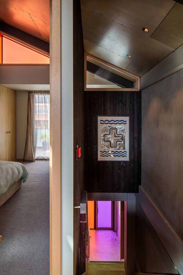A narrow interior hallway and stairway with dark vertical timber wall panelling, a warm wood-lined ceiling, recessed spotlights, and a textured grey wall. A framed artwork is mounted above the stair opening, while vivid purple and orange lighting glows from the lower level. A bedroom with carpet, curtains, and a bed is visible through the open doorway on the left.