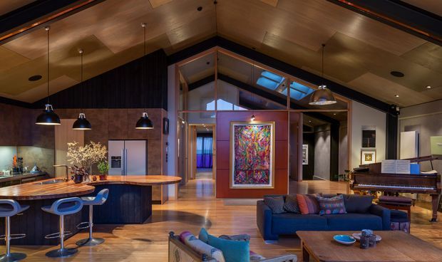 Open-plan contemporary living and kitchen interior with a dramatic vaulted timber ceiling, dark wood cabinetry and wall finishes, a curved kitchen island with bar stools, timber flooring, a blue sectional sofa, artwork, pendant lighting, and a piano. A glazed internal partition and clerestory glazing add visual openness and natural light.