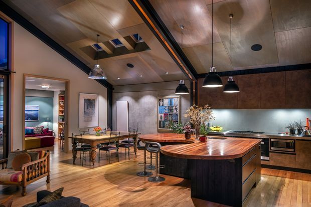 A warm, open-plan contemporary kitchen and dining area with extensive timber ceiling and flooring, dark cabinetry, a large curved-edge wood island, pendant lighting, and a stainless-steel backsplash. A connected living area is visible through the opening at left.