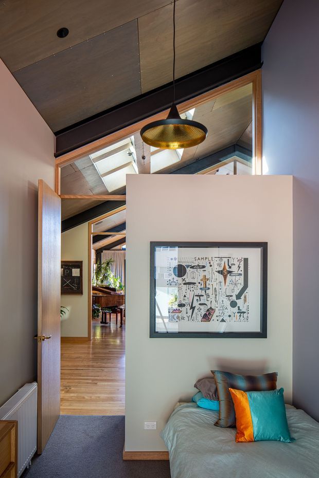 A compact bedroom viewed from the doorway, featuring a bed with layered teal, orange and brown cushions, a framed graphic artwork, and a warm timber door and floor. The room has a dramatic angled timber-lined ceiling with dark exposed structural beams, a black and brass pendant light, and a large internal glazed opening or mirror reflecting skylights and adjoining living areas. A hallway and open-plan space are visible beyond the door.