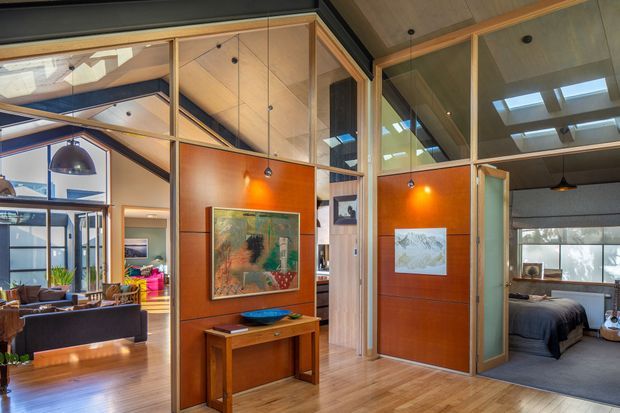 Interior view of a warm, open-plan modern home with extensive timber-framed glazing and internal partitions. A timber-floored living area with sofa and pendant lighting is visible to the left, while a bedroom with a bed is visible through a glazed doorway on the right. The central circulation zone features rich orange-brown timber wall panelling, built-in joinery, artwork, and high glazed sections with skylights.