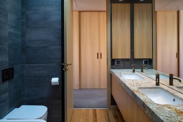 Contemporary bathroom with a double vanity, pale timber cabinetry, large framed mirrors, and a thick natural-stone benchtop. A separate toilet area is visible at left, finished with dark blue-grey wall tiles and a wall-mounted flush plate. Timber flooring and warm, integrated lighting create a refined, modern interior.