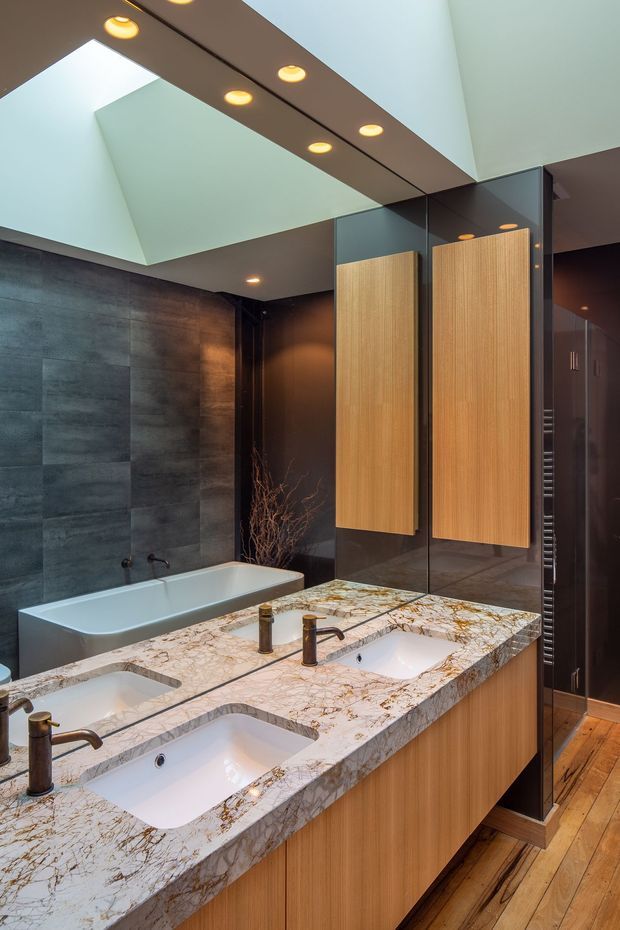 A luxurious contemporary bathroom with a double vanity, freestanding bathtub, large mirror panels, dark stone wall tiles, warm timber cabinetry, and a dramatic veined stone benchtop. A high ceiling with recessed warm lighting and a large skylight-like opening creates an airy yet moody atmosphere.