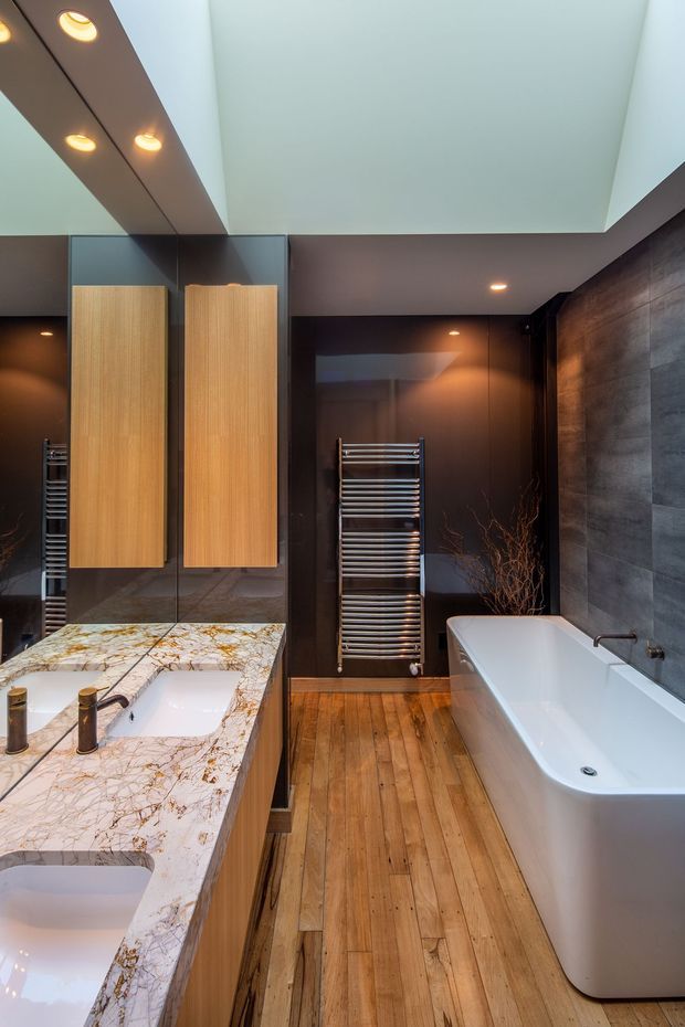 A luxurious contemporary bathroom featuring a freestanding white bathtub, double vanity with dramatic veined stone benchtop, mirrored cabinetry, dark glossy wall panels, timber flooring, a chrome heated towel rail, and recessed ceiling lighting. The room has a warm, moody, high-contrast atmosphere.