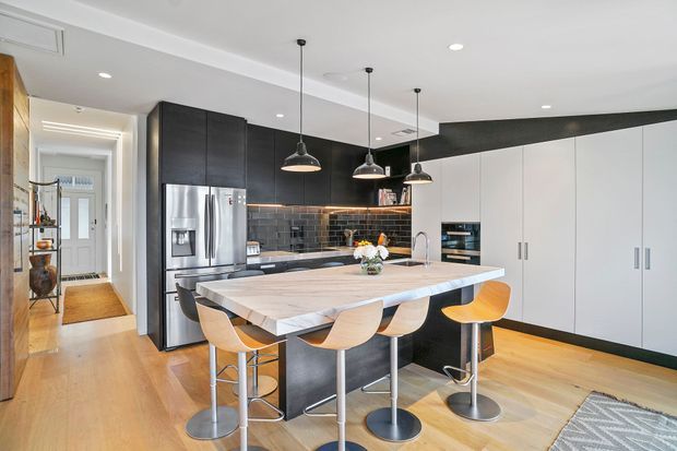 Contemporary open-plan kitchen with a large light stone island and integrated seating for four. The space features dark timber cabinetry, white full-height cabinetry, stainless-steel appliances, a dark tiled splashback, timber flooring, pendant lighting and a hallway visible beyond.