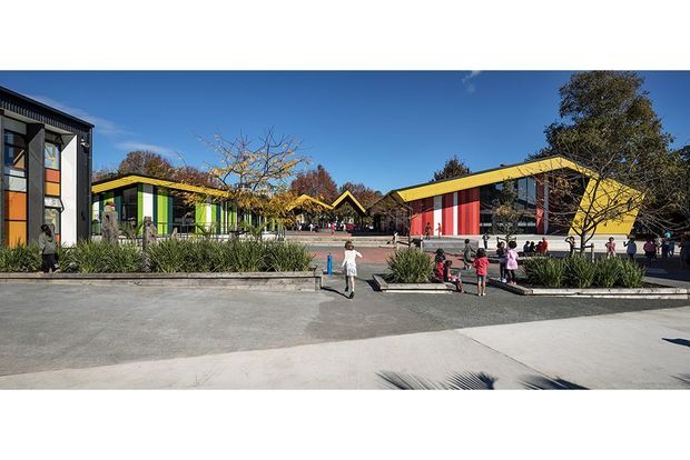 Wide exterior view of a contemporary, low-rise educational or commercial complex arranged around a paved courtyard. Several buildings feature bold yellow roof trims, dark grey façades, and brightly coloured vertical wall panels in orange, green, and red. Landscaping with raised concrete planters and grasses separates the buildings from the open pedestrian forecourt. Clear blue sky and people provide scale.