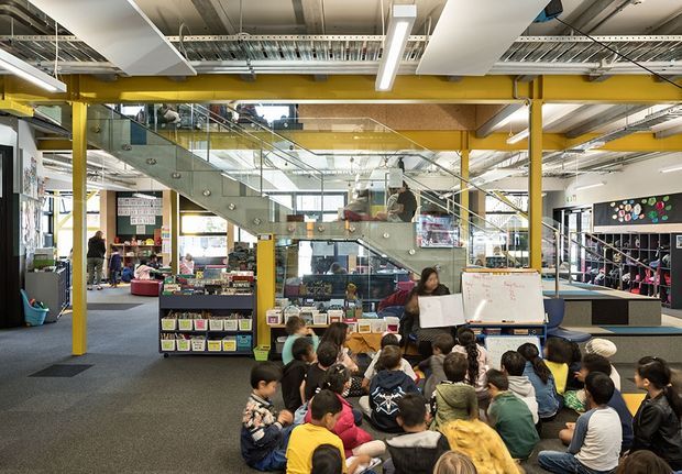 A bright, open-plan educational or commercial interior with exposed services, yellow structural columns and beams, a glass balustraded stair, library shelving, work tables, and a group of children gathered for a learning activity. The space has an industrial, adaptable learning-environment character with extensive glazing and visible upper-level circulation.