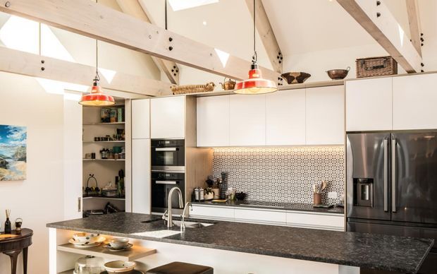 Bright contemporary kitchen with white full-height cabinetry, a large dark stone island, stainless-steel French-door refrigerator, integrated appliances, patterned tiled splashback, and open shelving. Exposed pale timber beams and a vaulted ceiling create a warm barn-like structural character, with red pendant lights adding colour.