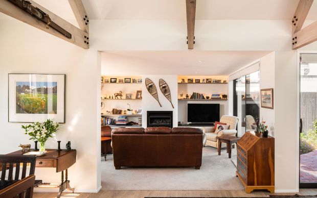Warm, light-filled living room viewed from an adjacent dining area, with exposed timber ceiling beams, white walls, built-in illuminated shelving, a fireplace, television, leather sofa, armchairs, and timber furniture. Large glazing on the right opens the room toward an outdoor area.