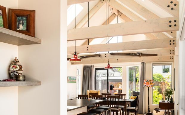 Bright open-plan dining area with a large timber dining table and chairs beneath a dramatic high gabled ceiling. Exposed pale timber beams and structural connectors dominate the architecture, with extensive glazing and doors opening toward a leafy outdoor area. Red pendant lights, floating display shelves, framed artwork, and decorative objects add warmth and character.