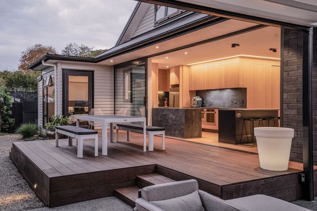 Exterior view of a contemporary home with a covered, open-sided outdoor living area and integrated kitchen opening onto a raised timber deck. The space includes a built-in barbecue and cabinetry, stone-look island, dining table with bench seating, and lounge seating in the foreground. The house features light weatherboard cladding, dark brick accents, broad roof eaves, and warm integrated lighting.