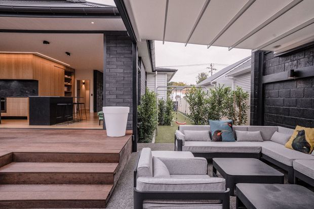 A contemporary outdoor entertaining area with a large light-grey sectional lounge and black coffee tables beneath a covered pergola. The space adjoins an open-plan kitchen with warm timber cabinetry, a dark island and black brick feature walls. Broad timber steps connect the elevated interior to the paved outdoor seating area, with planted screening along the boundary.
