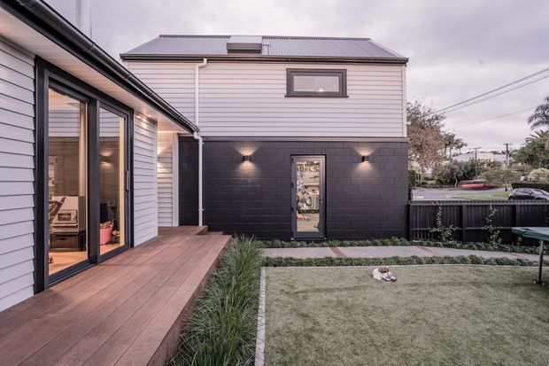 A contemporary residential courtyard viewed from a timber deck. The two-storey building combines pale horizontal weatherboard cladding above a dark painted blockwork base, with black-framed glazing, exterior wall lights, and a small landscaped lawn bordered by ornamental grasses. A hot tub and neighbouring residential context are visible beyond the courtyard fence.