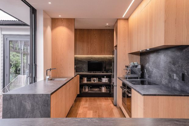A compact contemporary galley kitchen with warm timber cabinetry, dark stone benchtops and splashback, integrated appliances, a central island, and a narrow living or media area at the rear. Full-height glazing opens to an outdoor area on the left, bringing in natural light.