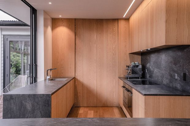 A compact, contemporary kitchen with warm timber cabinetry and full-height timber wall panelling. Dark stone benchtops and matching splashback create contrast, with integrated appliances, a sink, island bench, and large glazed doors opening toward an exterior space. Recessed spotlights and a linear LED add modern illumination.