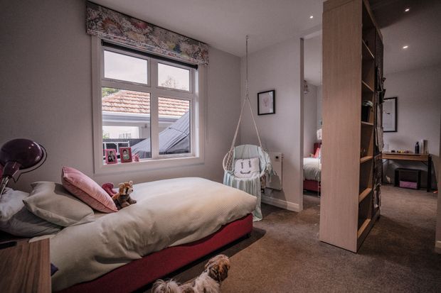 A warm, compact bedroom with a low upholstered bed, layered neutral bedding and pink accents, a hanging egg chair, carpeted floor, framed artwork, and a large timber open-shelving partition separating the sleeping area from an adjoining workspace or circulation area. A second bed is partially visible through the opening behind the partition.