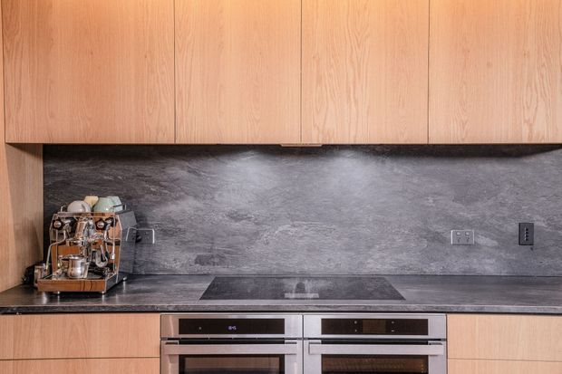 Contemporary kitchen detail featuring warm wood cabinetry, a dark grey stone-look splashback and benchtop, integrated double ovens, and a coffee machine. The composition focuses on the built-in cabinetry and cooking zone.
