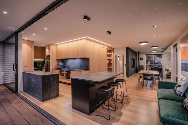 Open-plan contemporary kitchen and dining area viewed from an adjoining outdoor deck through large sliding glazing. The room features extensive warm timber cabinetry, dark stone island benchtops, integrated linear lighting, black-framed glazing, a dining table, and a green upholstered sofa.