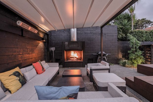 A covered outdoor lounge with a large sectional sofa, armchairs, coffee tables and an illuminated built-in fireplace. The space has dark painted masonry walls, a slatted retractable-style roof canopy, integrated outdoor heating, and gravel flooring, with planted greenery visible around the perimeter.
