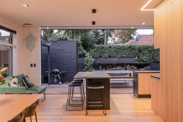A warm contemporary open-plan kitchen and dining area opening onto a landscaped outdoor entertaining space. The room features light timber flooring and cabinetry, a dark stone kitchen island with stools, a timber dining table, and an adjacent green sofa seating area. Large glazed openings provide a strong indoor-outdoor connection to a paved terrace with a barbecue, bench seating, dark masonry walls, hedging, and established planting.