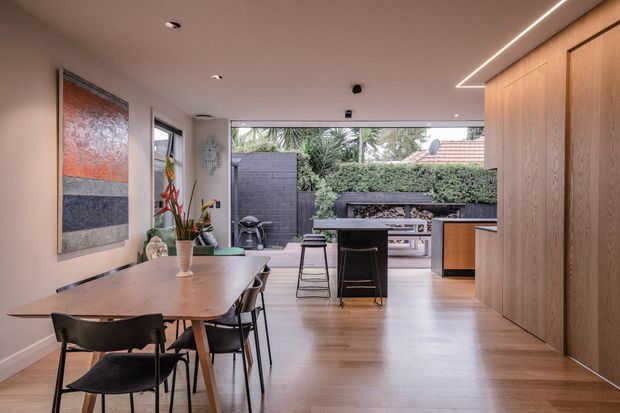 A contemporary open-plan dining and kitchen area with pale timber flooring, a long timber dining table and black chairs, full-height timber cabinetry, and a compact kitchen opening directly onto a landscaped outdoor entertaining area. Linear recessed lighting, simple white walls, and expansive glazing create a bright, refined connection between indoors and outdoors.