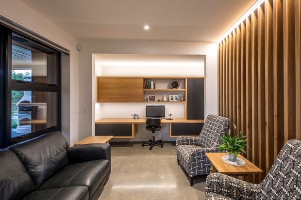 A compact, warm-toned home office and sitting area. A built-in timber desk and shelving unit is set into a recessed wall niche, with a computer, office chair and integrated cabinetry. Black leather seating and patterned armchairs occupy the foreground, while a full-height vertical timber batten feature wall with concealed linear lighting defines the right side. A dark-framed window is visible on the left, and the polished concrete floor and neutral walls create a contemporary, refined interior.