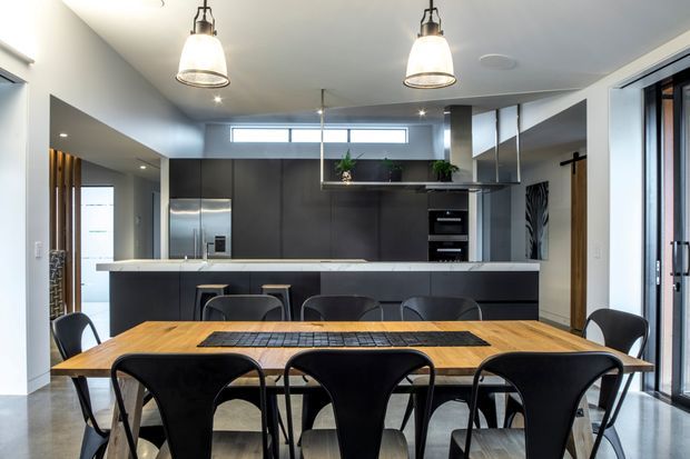 Contemporary open-plan kitchen and dining area with a long natural timber dining table and black chairs in the foreground. The kitchen features dark matte cabinetry, a pale stone island benchtop, integrated appliances, pendant lighting, stainless-steel refrigerator, open shelving, and large glazed doors at right. Neutral finishes and strong black-and-wood contrast create a refined, functional entertaining space.