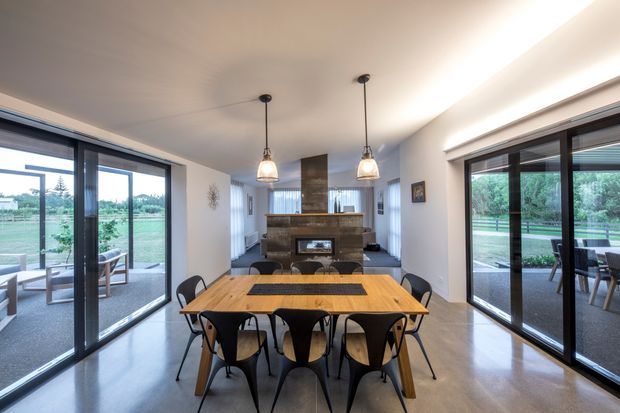 Open-plan dining and living area with a large timber dining table and black sculptural chairs in the foreground. Floor-to-ceiling dark-framed glazing and sliding doors open toward an outdoor setting on both sides. A central fireplace with a stone or brick surround anchors the rear living zone, beneath two pendant lights and a high, sloping ceiling.