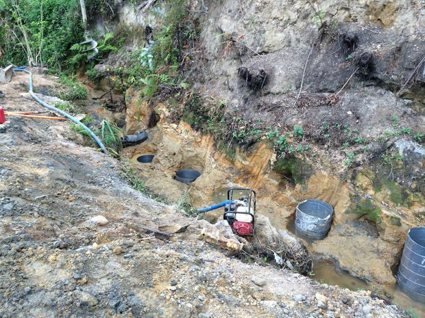 A rugged excavated outdoor site with exposed soil and rock, drainage channels, hoses, buckets, and a small portable pump. Vegetation grows along the cut hillside, suggesting active earthworks or site drainage and construction work.