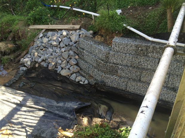 A small landscaped drainage or erosion-control area with stacked gabion baskets filled with grey stone, a rock-lined channel, black geotextile sheeting, muddy water, and exposed white utility pipes. Dense vegetation surrounds the rough outdoor construction feature.