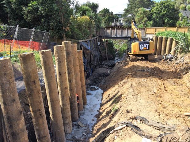 Construction site showing a deep excavated trench with temporary timber shoring piles and retaining supports. A CAT excavator is working at the edge of the excavation, with fencing, soil stockpiles, vegetation, and a pedestrian bridge visible in the background.