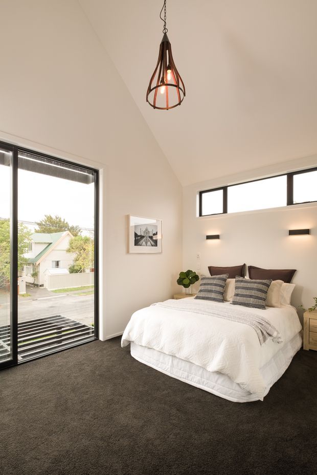 Bright contemporary bedroom with a large upholstered bed dressed in white bedding and layered grey striped cushions, dark carpet, pale walls, black-framed glazing, minimalist wall sconces, and a sculptural hanging pendant light. A framed monochrome artwork and small bedside tables with plants complete the room. The large window provides views toward neighbouring residential buildings.