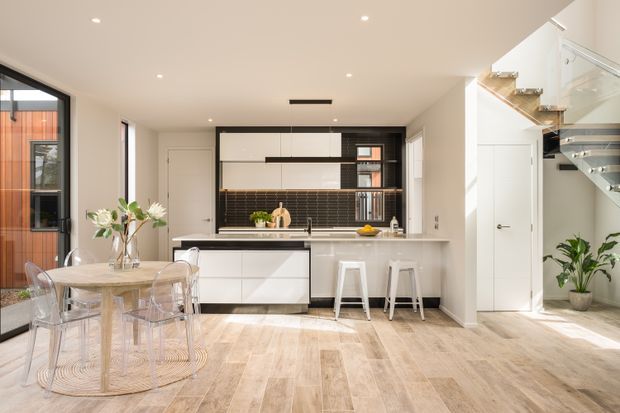 Bright open-plan kitchen and dining area with white flat-panel cabinetry, a long island with seating, dark tiled splashback, pale timber-look flooring, and a glass dining table with transparent chairs. A glazed stairway and landing are visible to the right, with large windows and doors bringing in natural light.