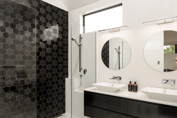 Contemporary bathroom featuring a walk-in shower lined with dark hexagonal tiles, a frameless glass partition, and a double floating vanity with white vessel basins, wall-mounted chrome taps, round mirrors, and black cabinetry. Bright natural light enters through a high window.