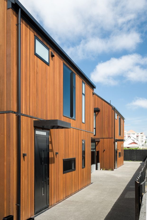Exterior view of a narrow multi-storey contemporary residential building or row of attached dwellings, clad in warm vertical timber boards with dark metal window and door frames. The facade has a simple angular roofline, tall narrow windows, projecting dark entry canopies, and a concrete pedestrian access path. A suburban neighbourhood is visible beyond.