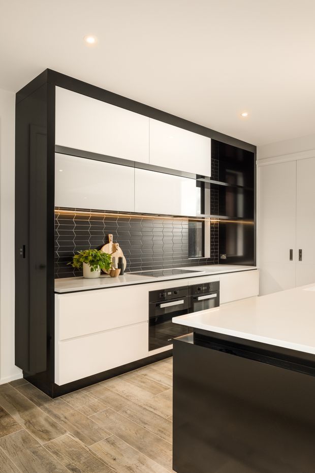 A contemporary, minimalist kitchen with a full-height black-framed wall of white handleless cabinetry, integrated black appliances, and a dark hexagonal tile splashback. Warm under-cabinet lighting highlights the benchtop and decorative timber and plant accents. A large white island with a black base occupies the foreground, and the room has timber-look flooring and recessed ceiling lights.