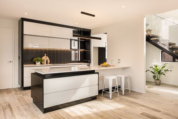 Bright contemporary open-plan kitchen with a large white island, black-framed and black-accented cabinetry, dark horizontal subway-tile splashback, pale timber-look flooring, and a white breakfast counter with stools. A glazed stairway with dark handrails is visible to the right, reinforcing the open-plan layout.
