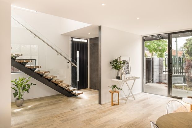 Bright contemporary open-plan entry and living area featuring a floating timber staircase with black metal supports and glass balustrade, pale timber flooring, a dark pivot-style entry door, white walls, and full-height glazed doors opening to a small courtyard. Minimal furniture and indoor plants create an airy, refined interior.