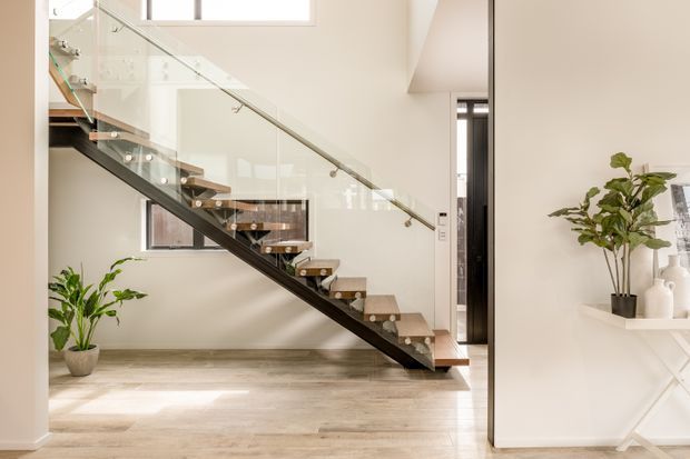 Bright contemporary interior view focused on an open-riser staircase with timber treads, dark structural stringers, and clear glass balustrades. The staircase rises beside a light neutral wall with high windows, pale timber-look flooring, and minimal styling including potted plants and a white console.