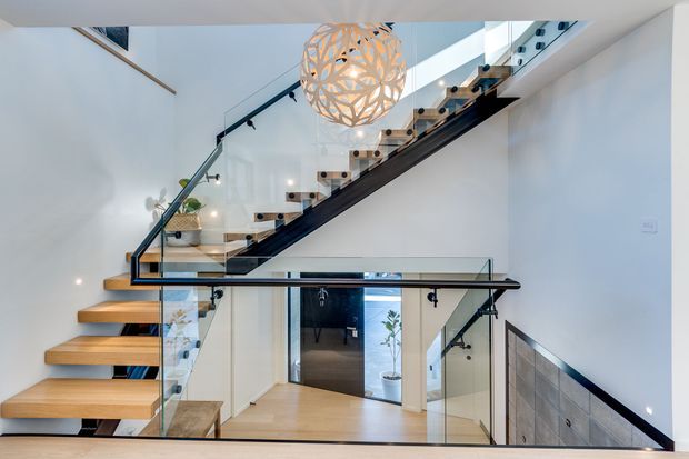 Bright contemporary residential stairway with floating timber treads, black structural stringers and handrails, clear glass balustrades, and a sculptural spherical pendant light. The stair connects multiple levels around a compact central landing, with white walls and pale timber flooring creating an airy, refined interior.