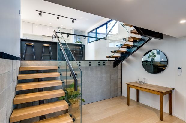 Bright contemporary interior showing a split-level entry and stair hall. Floating timber stairs with black metal stringers and clear glass balustrades lead upward beside a compact kitchen or bar area with dark cabinetry and stools. A second floating stair flight rises overhead, while a tiled half-height wall, timber console table, round mirror, white cabinetry, and large windows complete the space.